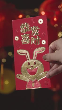 Vertical-Video-Of-Woman-Holding-Card-Celebrating-2023-Chinese-New-Year-With-Chinese-Lanterns-Hung-In-Background